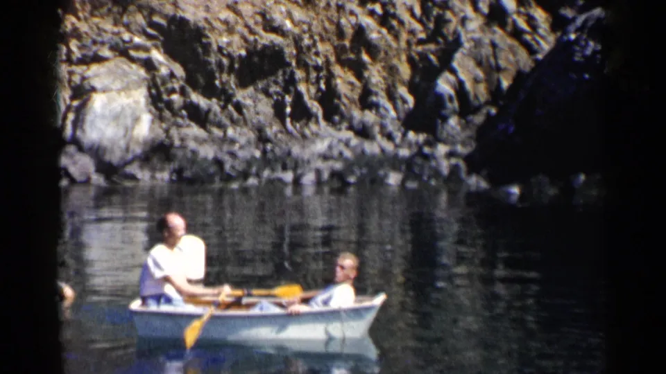 1955 California 8mm frame showing two men in a white rowboat with yellow oars near shaded rocks and reflective water.