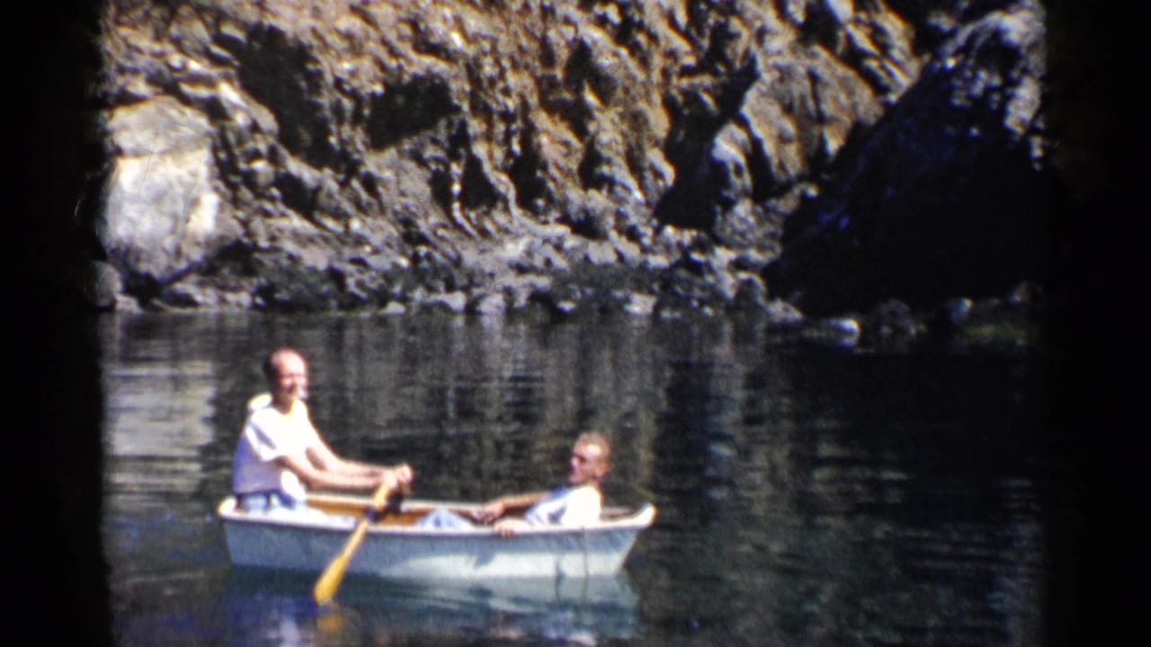 1955 California home-movie frame of two adult men in a small white rowboat near a rocky shoreline on still water.