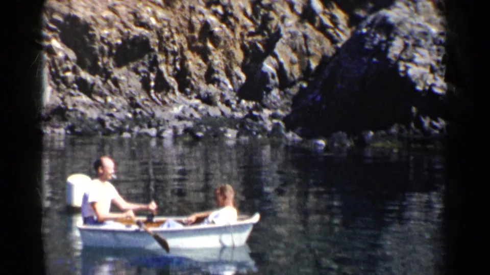 Two adult men appear in a small white rowboat on calm water, with one handling an oar near a rocky California shoreline.