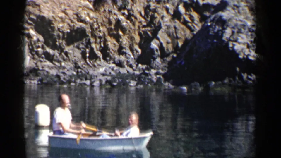 A white rowboat carrying two adult men moves across calm water near a white buoy and rugged rocks in archival 8mm footage.
