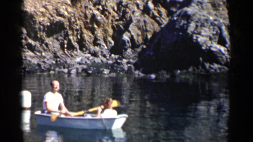 Two adult men continue across the water in a white rowboat, framed by rocky cliffs and 8mm film borders.