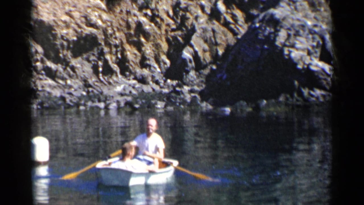 Man and child in a small rowboat on calm water near rocky cliffs.