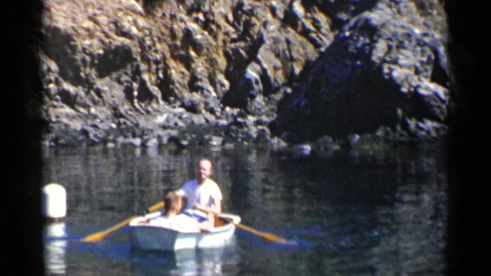 1955 California home-movie frame showing a man and child in a small rowboat on a quarry-like lake beside steep rock walls.