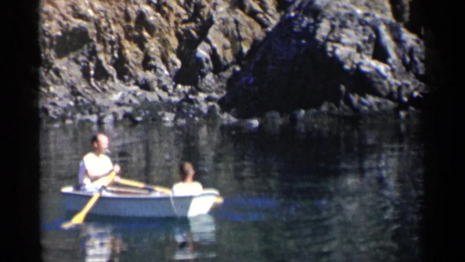 1955 California home-movie frame with two people in a small rowboat crossing still water beneath a steep rocky quarry wall.