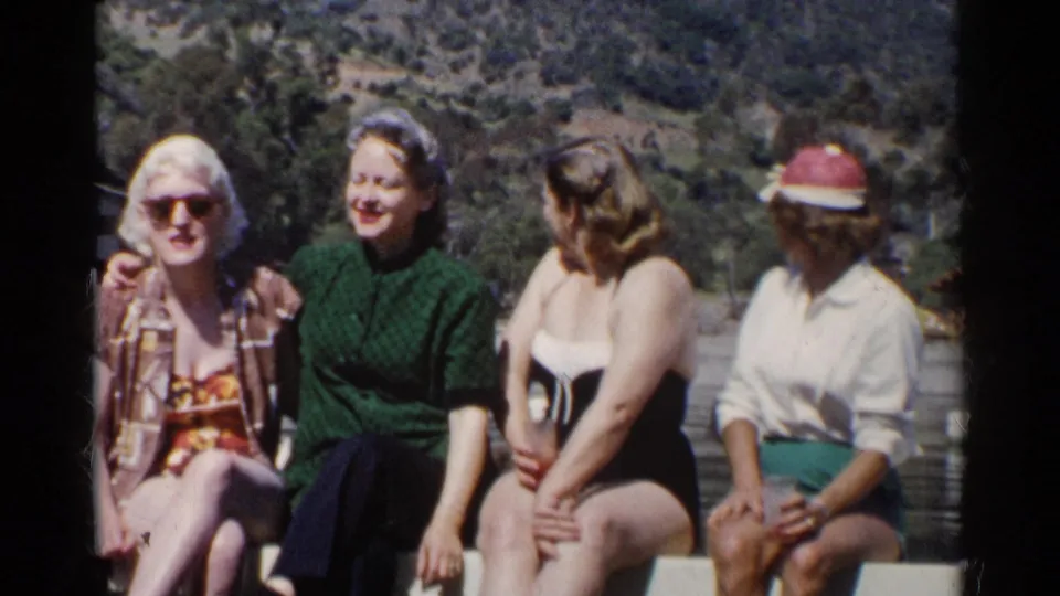 1955 California home-movie frame showing four women on a dock by a lake, wearing swimsuits, sunglasses, and casual summer clothing.