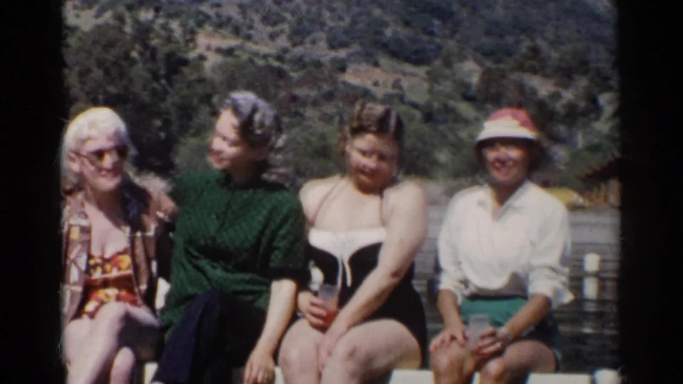 1955 California home-movie frame of four women smiling on a dock beside a quarry-like lake, with period swimwear and summer clothing visible.