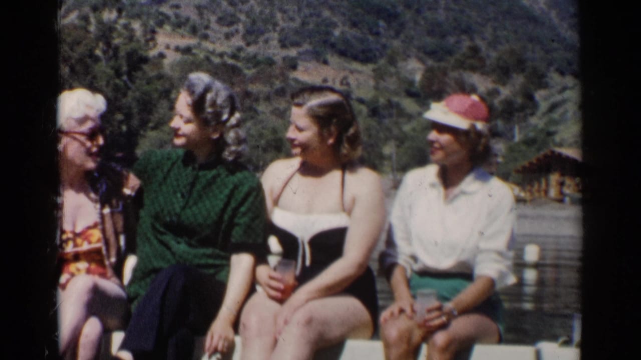 Four women sit on a railing by a lake, smiling and talking in bright sun.