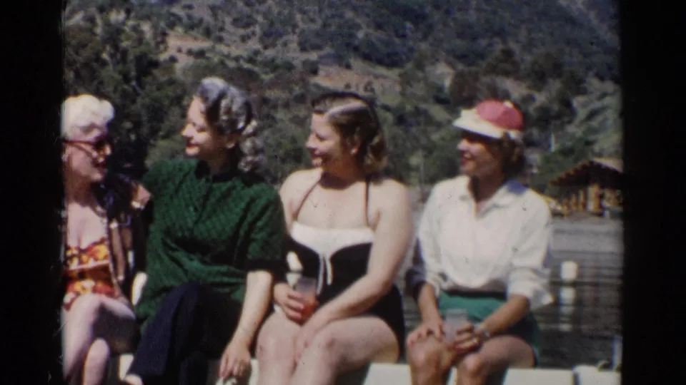 Four women in 1950s California lake footage sit together on a railing, wearing swimwear and casual summer clothing.