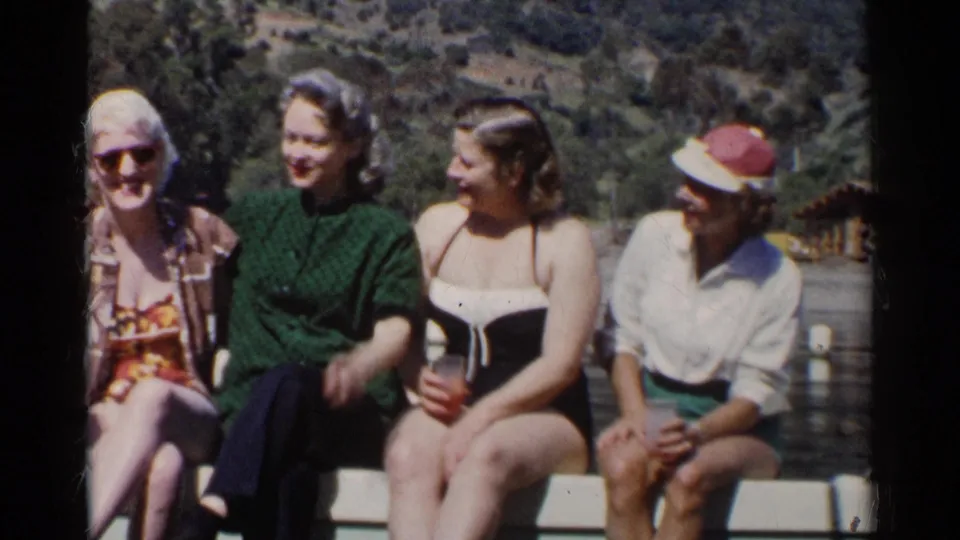 Four women sit on a lakeside railing in 1950s California, with one older woman looking toward the camera.