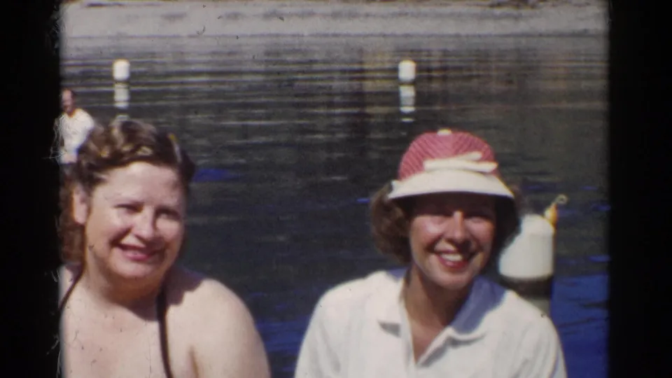 1955 California frame showing two women smiling near a pond, one in a swim top and one in a white shirt with a visor.