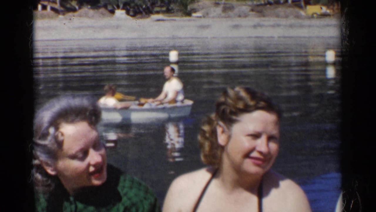 Two women near a calm lake with a rowboat passing behind them.