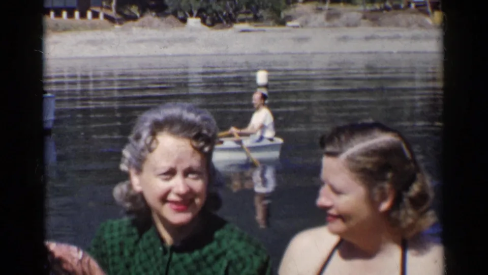 Two women smile and talk near a California lake, with a man rowing a small boat and another passenger visible in the background.
