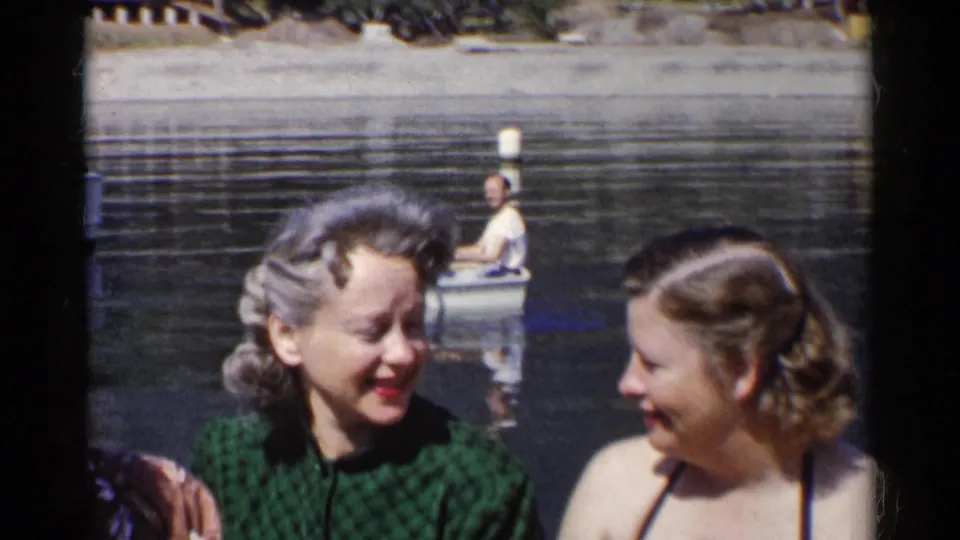 A close home-movie view shows two women conversing by calm water, with a rowboat, oars, and distant shore visible behind them.