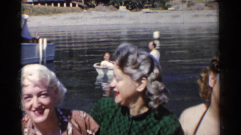 Three women appear at the edge of frame near a calm California lake, with a small rowboat and rower visible behind them.
