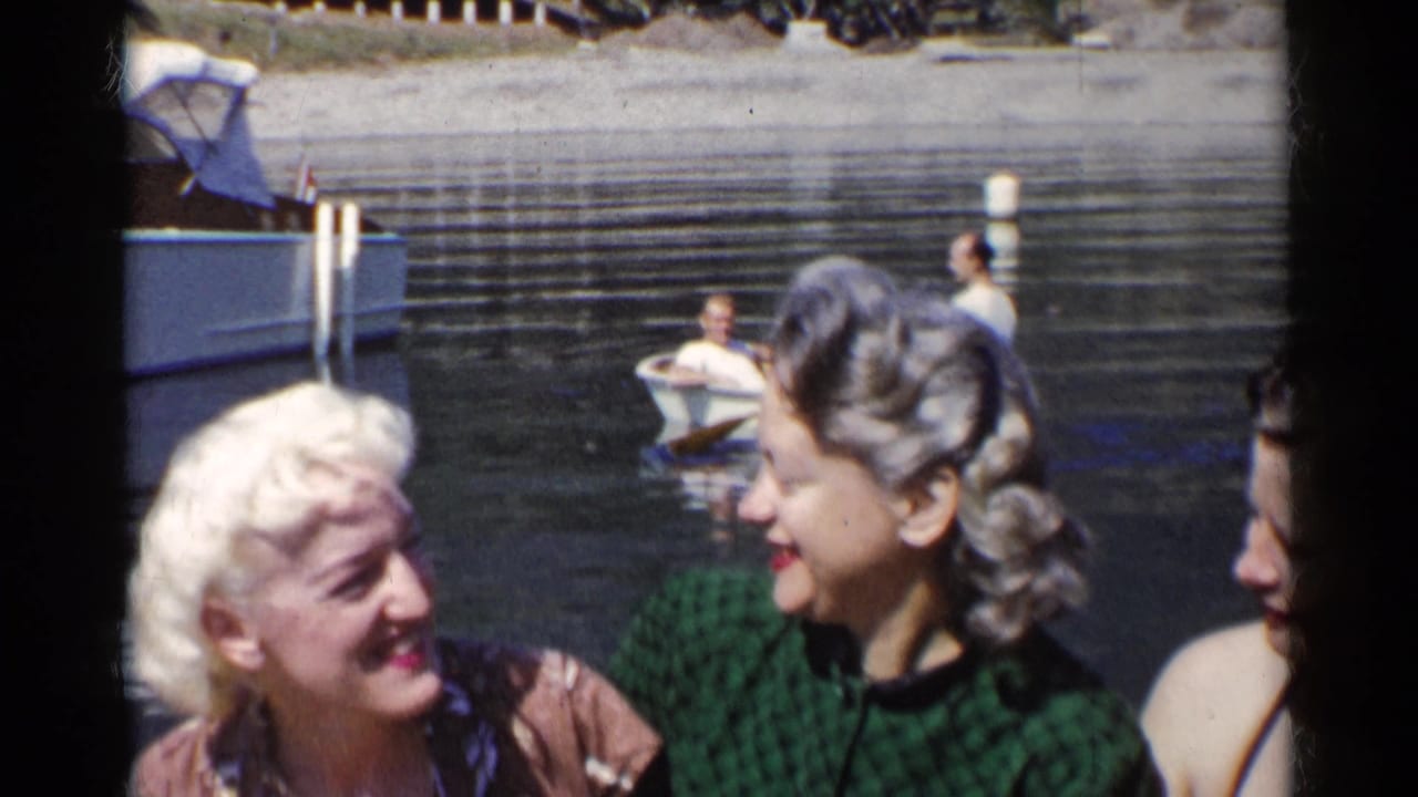 Three young women laugh close to the camera beside river water and boats.