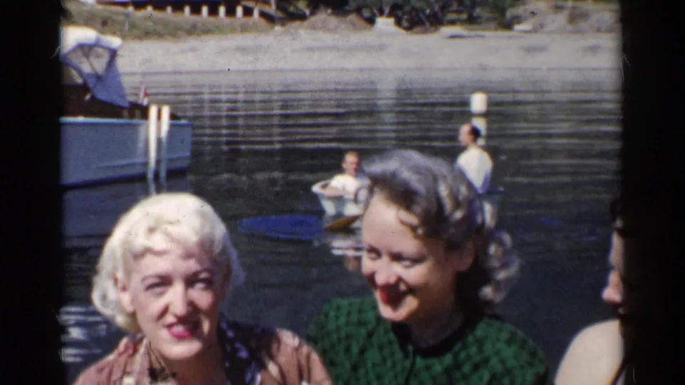 Two young women smile near the camera in California, with calm river water, a small rowboat, and a docked boat visible behind them.