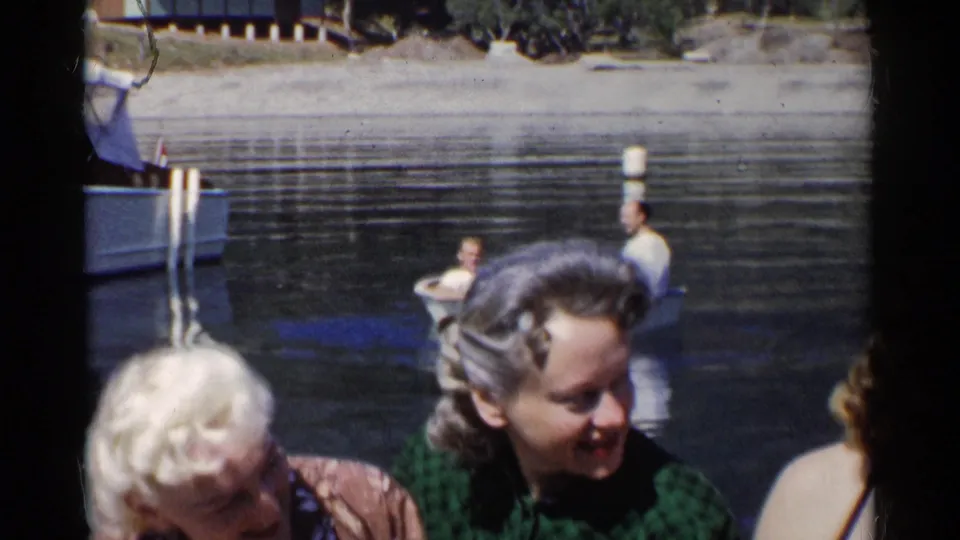 The home-movie frame shows young women lowering their gaze and smiling beside the river, with a rowboat and shore in the background.