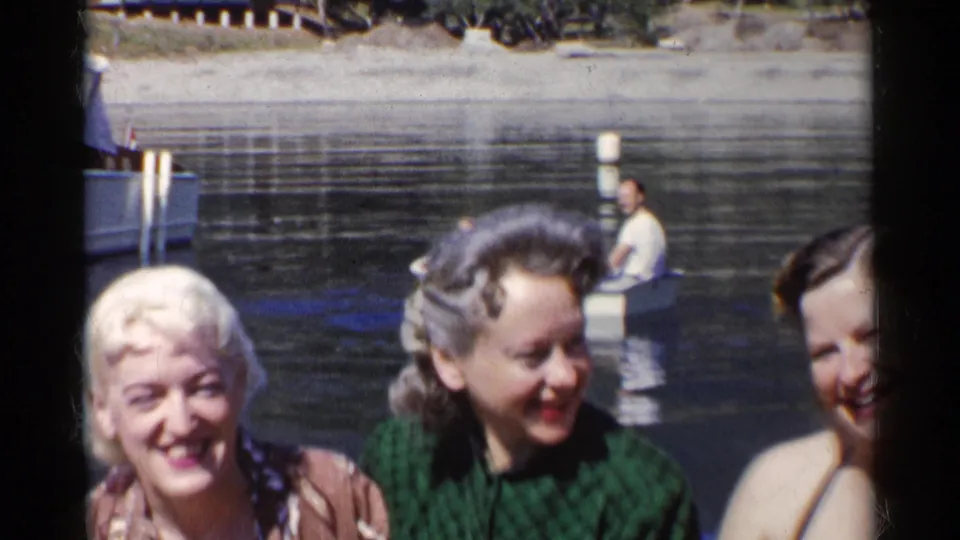 Three young women in 1955 California smile and laugh by the river as two people in a small rowboat appear in the background.