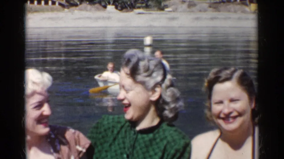 1955 California home-movie frame showing women near the camera as a small rowboat moves on a lake behind them.