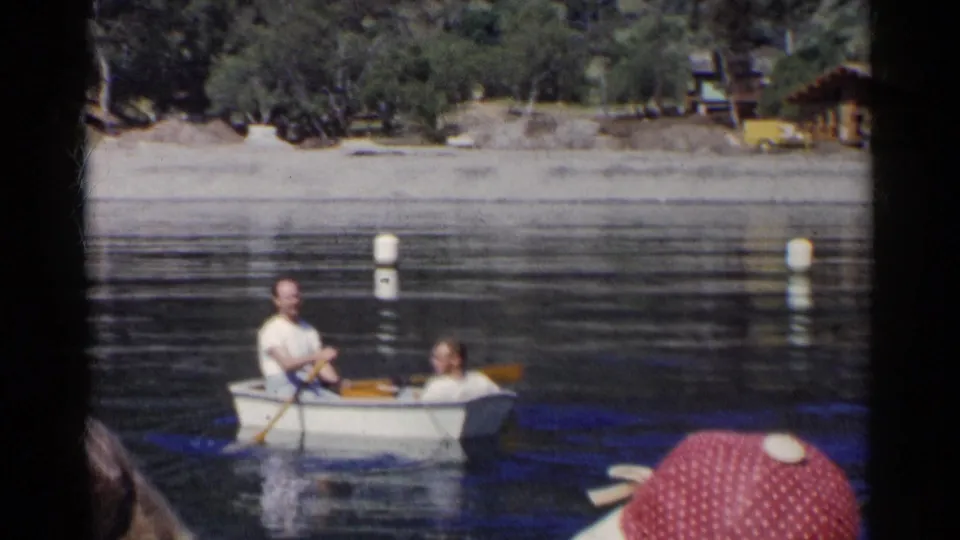 Two men in a small white rowboat move across a California lake, with sandy shore and trees behind them.