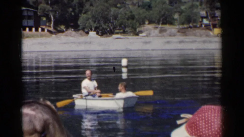 Home-movie frame of two men rowing a small boat on a California lake, with spectators partly visible in the foreground.