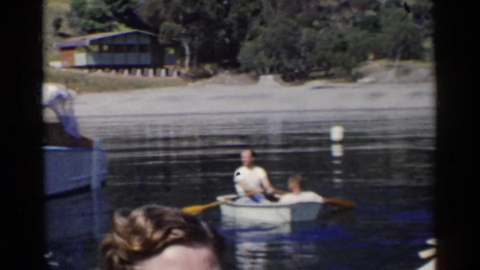 Two men row a small boat on a California lake as shoreline trees, a cabin, and spectators appear around the frame.