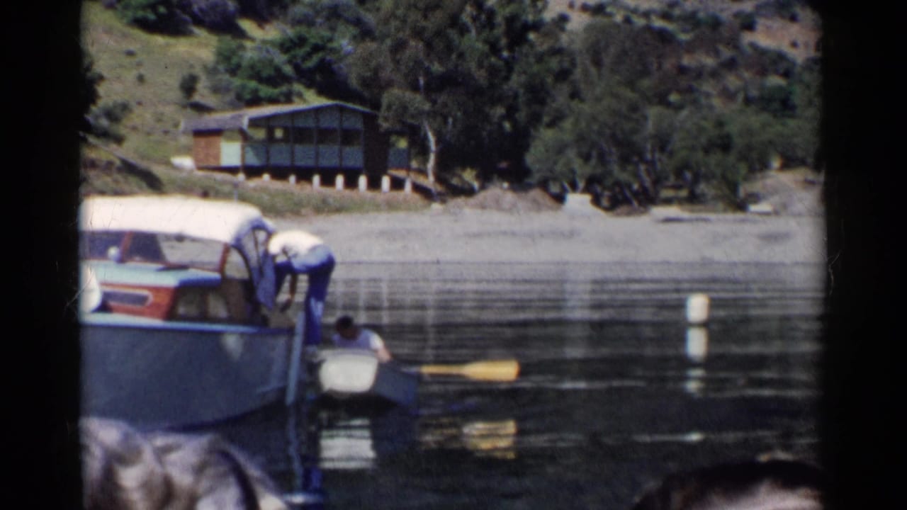 1955 California lake scene with a person in a rowboat beside a larger covered motorboat, with a hillside cabin and trees beyond the water.