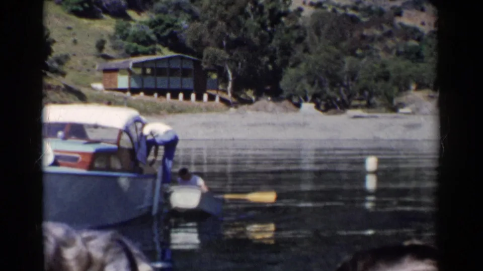 1955 California lake scene with a person in a rowboat beside a larger covered motorboat, with a hillside cabin and trees beyond the water.