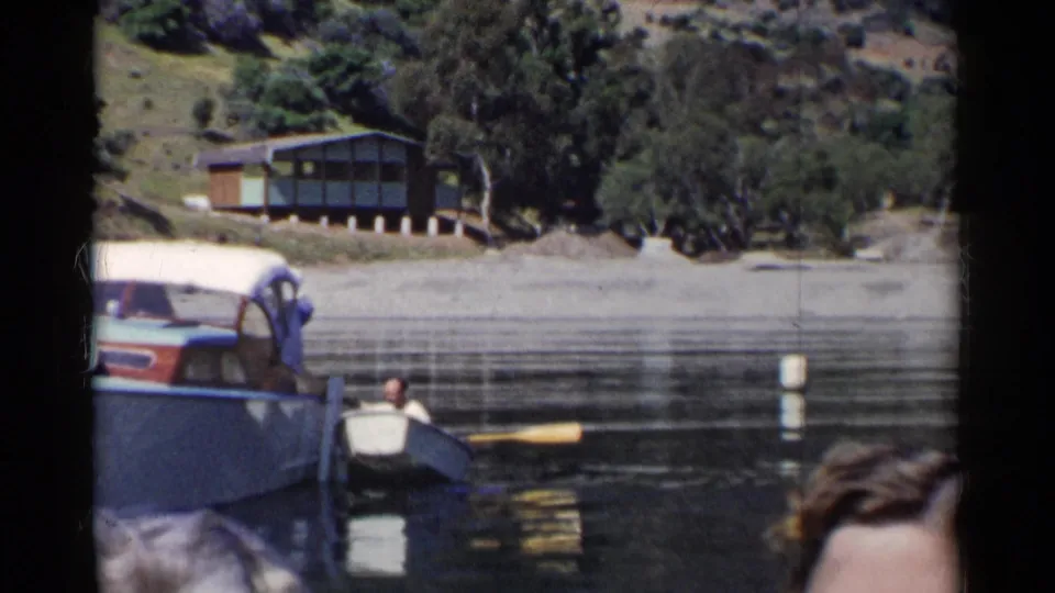A person rows or steadies a small boat near a larger covered boat on a California lake, with a cabin and trees in the background.