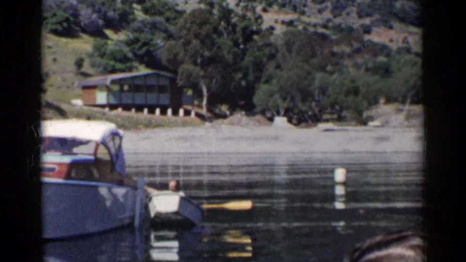 A small boat with a seated person and yellow oar sits beside a covered motorboat on a 1955 California lake, with a cabin above the shore.