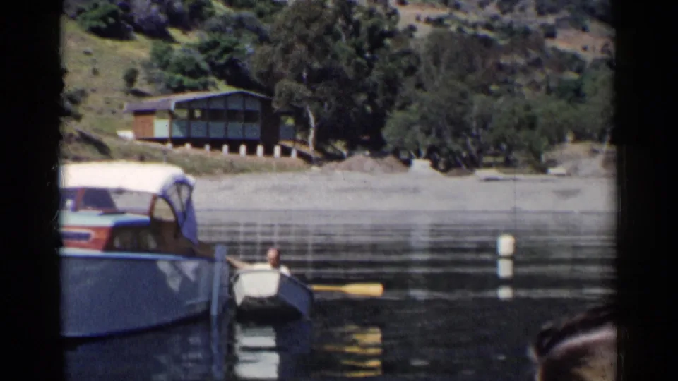 A rowboat with a visible passenger sits near a larger covered motorboat on a California lake, with a buoy, wooded shore, and cabin behind.
