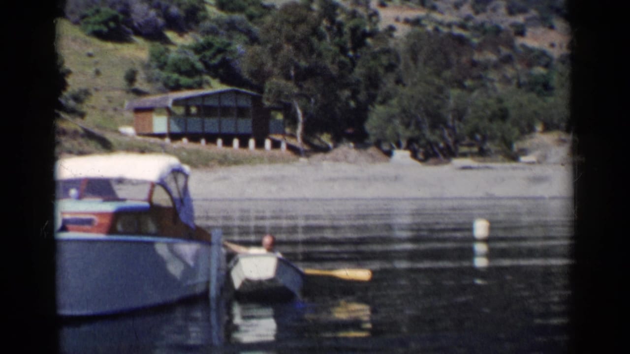 1955 California home-movie frame showing a docked motorboat, a small rowboat with a distant person, calm cove water, and a hillside shack.