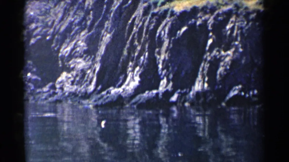 1955 California coastal frame focused on steep eroded rock formations beside still cove water, with reflections and 8mm gate edges visible.