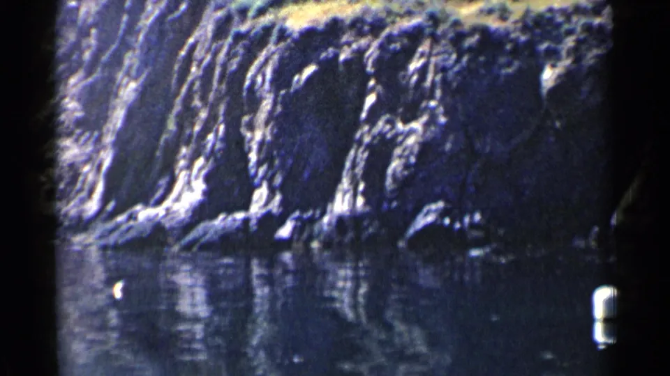 A 1955 California cove view shows textured rock faces, water reflections, brush above the cliff, and the soft grain of 8mm home-movie film.