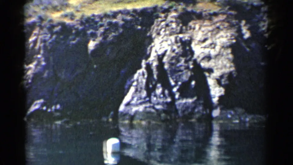 1955 California coastal frame with textured rocks, still water, dark shadows, and a white buoy giving scale in the foreground.