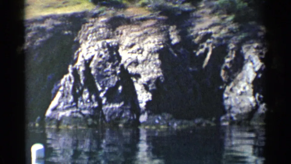 1955 California shoreline view showing a tall rock formation, still water, and a partly framed white buoy at the lower left.
