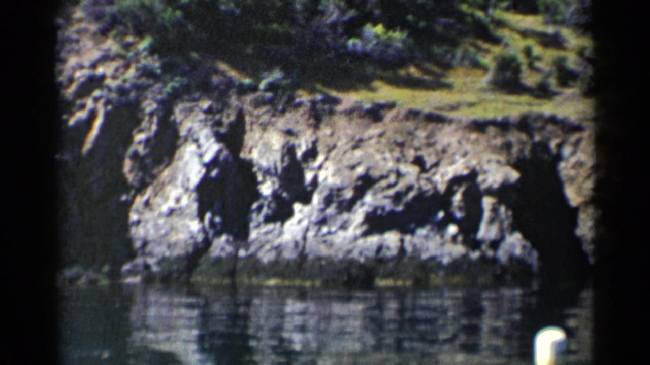 1955 California 8mm frame showing a rocky lake shoreline, green vegetation above the bank, and still water below.