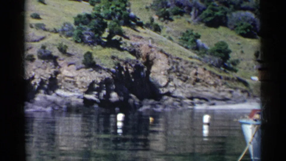 1955 California shoreline frame showing green hills, rocky banks, white buoys, calm water, and a partial small boat at the right edge.