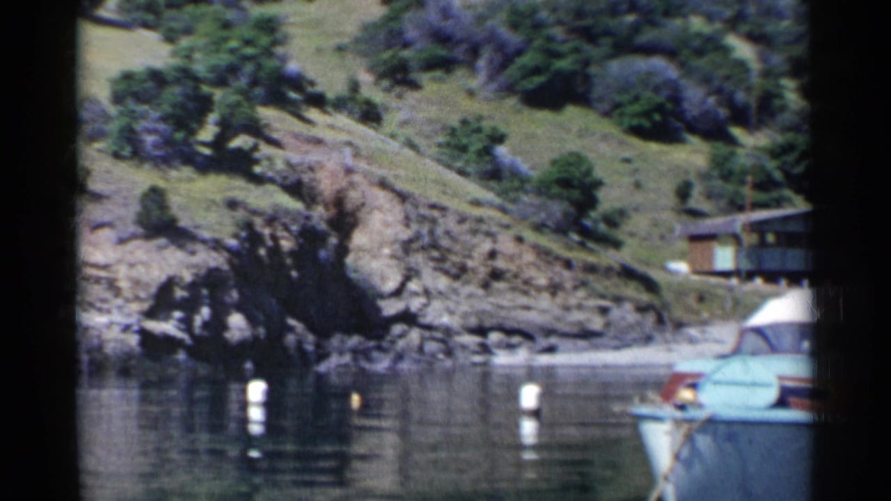 1955 California 8mm frame showing a blue motorboat partly in view beside calm cove water, white buoys, rocks, and a grassy hillside.