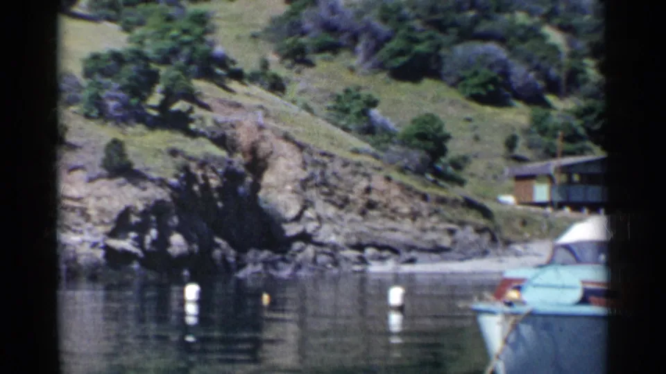 1955 California 8mm frame showing a blue motorboat partly in view beside calm cove water, white buoys, rocks, and a grassy hillside.