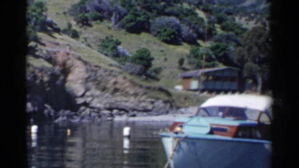 1955 California home-movie frame with a blue and white motorboat in a coastal cove, white buoys, rocks, hillside brush, and a small shore structure.