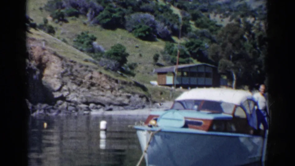 1955 California 8mm frame showing a person on a blue motorboat moored in calm cove water beneath a rocky, grassy hillside.