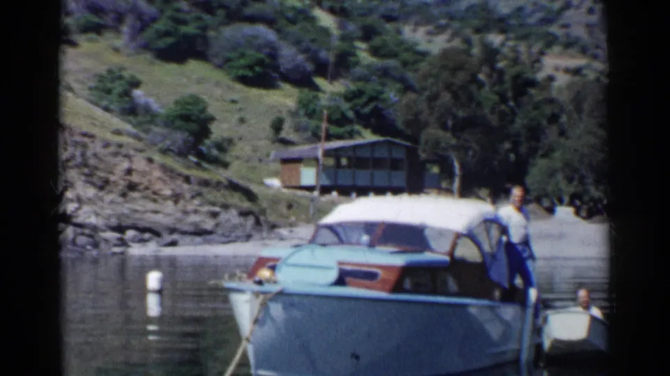 1955 California home-movie frame showing a person standing on a blue motorboat, with a small dinghy, buoys, calm water, rocks, and hillside behind.