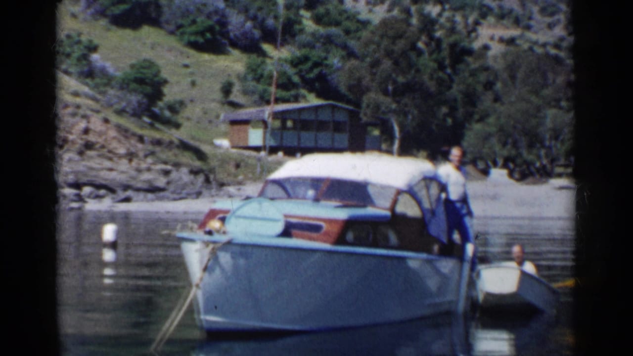 Man stands on a light-blue motorboat beside a small rowboat on a calm lake.