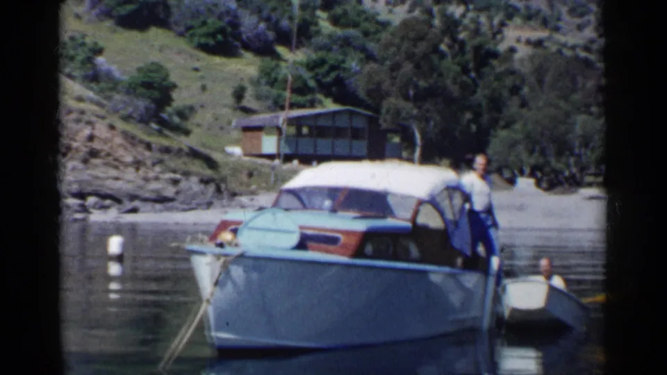 1955 California 8mm frame of a man on a light-blue motorboat near a small rowboat, with a cabin and wooded hillside behind.