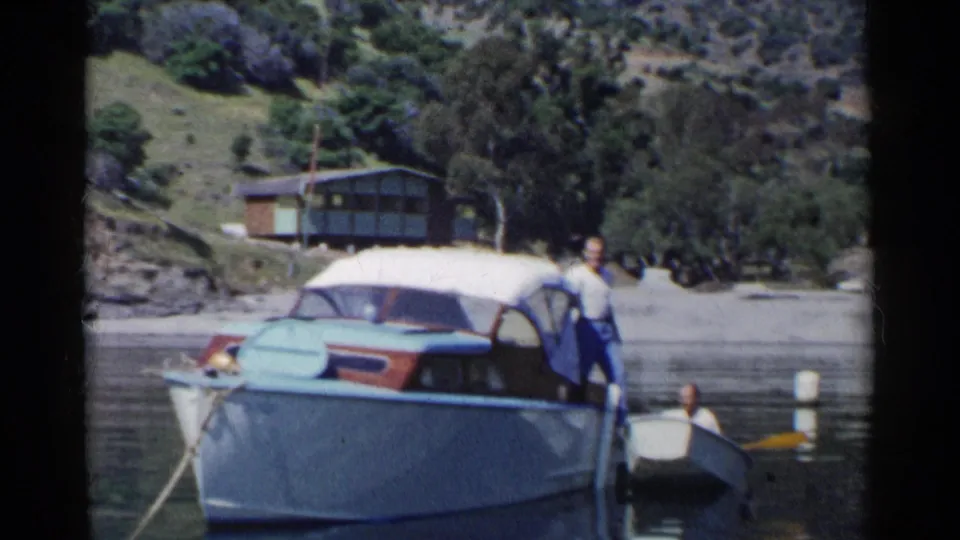 A man stands on the larger boat beside a small rowboat on a California lake, with shoreline trees and a cabin visible.