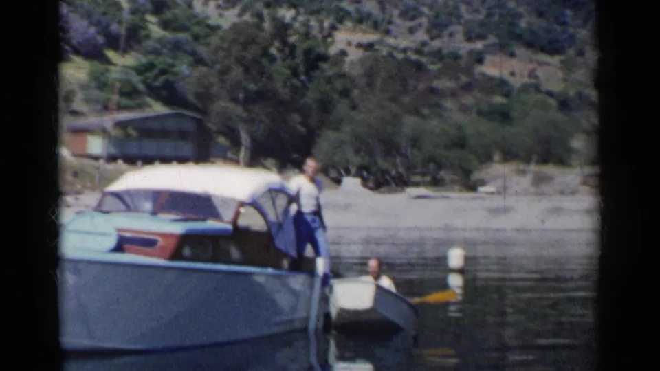 The 8mm frame shows the motorboat and adjacent rowboat near a wooded California shoreline, with the man positioned at the side of the larger boat.