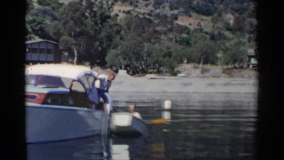 A man bends over from the larger motorboat toward a person or position in the rowboat, suggesting assistance during a 1955 California lake outing.