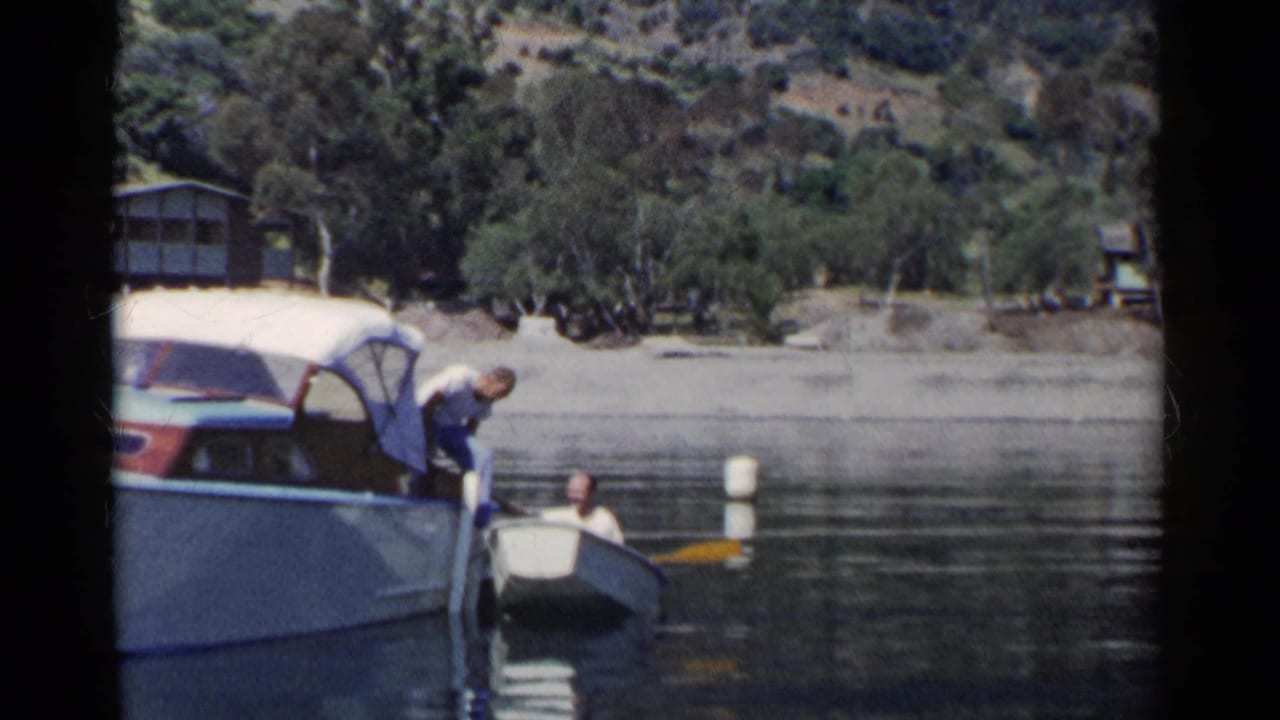 In 1955 California home-movie footage, a man transfers from a larger motorboat to a small rowboat near a wooded lakeshore.