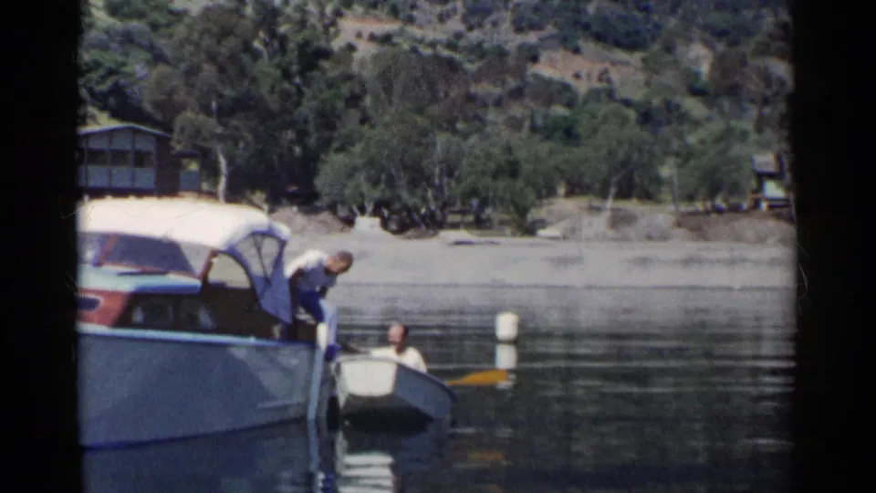 In 1955 California home-movie footage, a man transfers from a larger motorboat to a small rowboat near a wooded lakeshore.